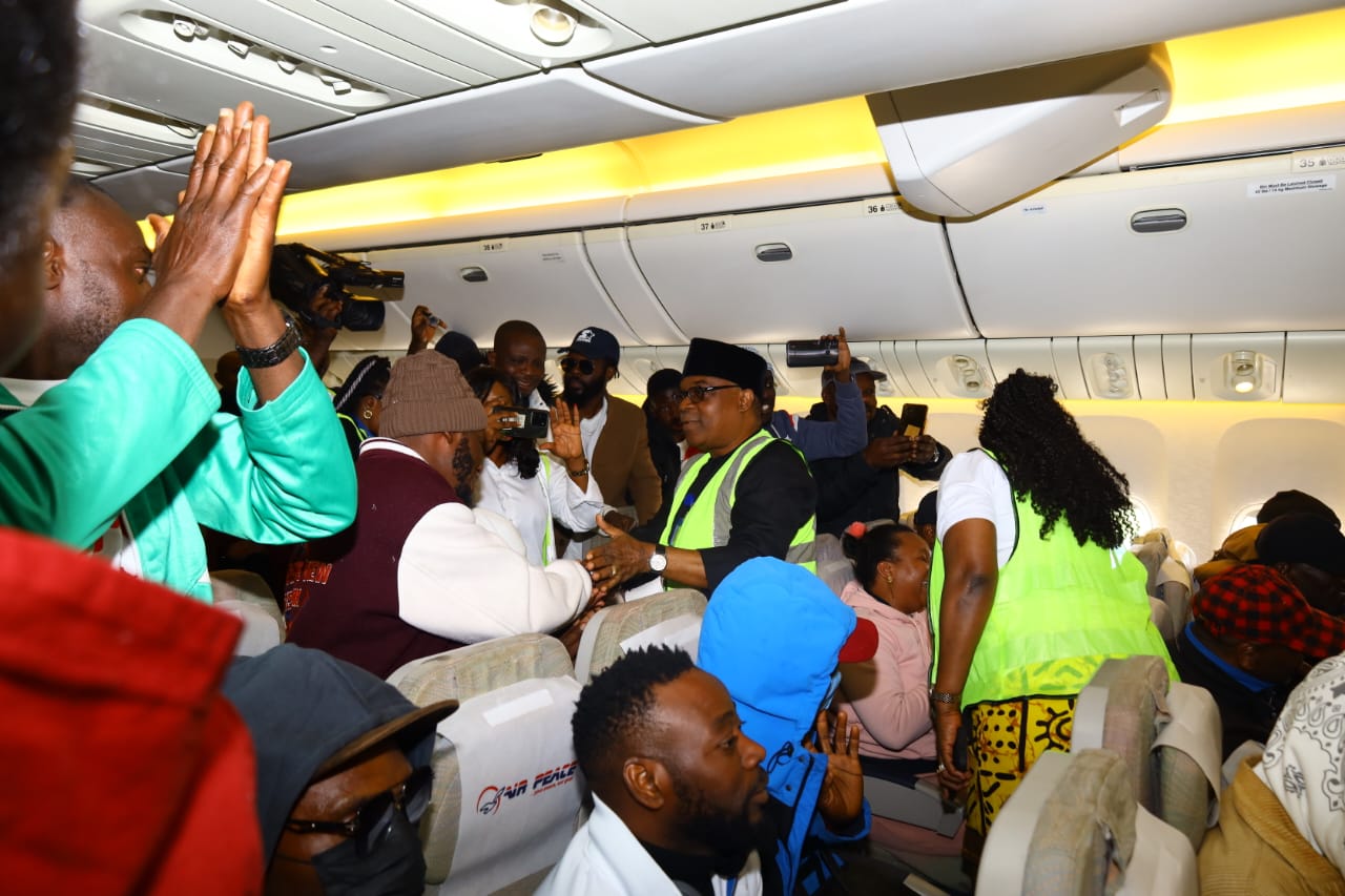Crowded airplane cabin with passengers standing in the aisle, a man in a safety vest greets others.