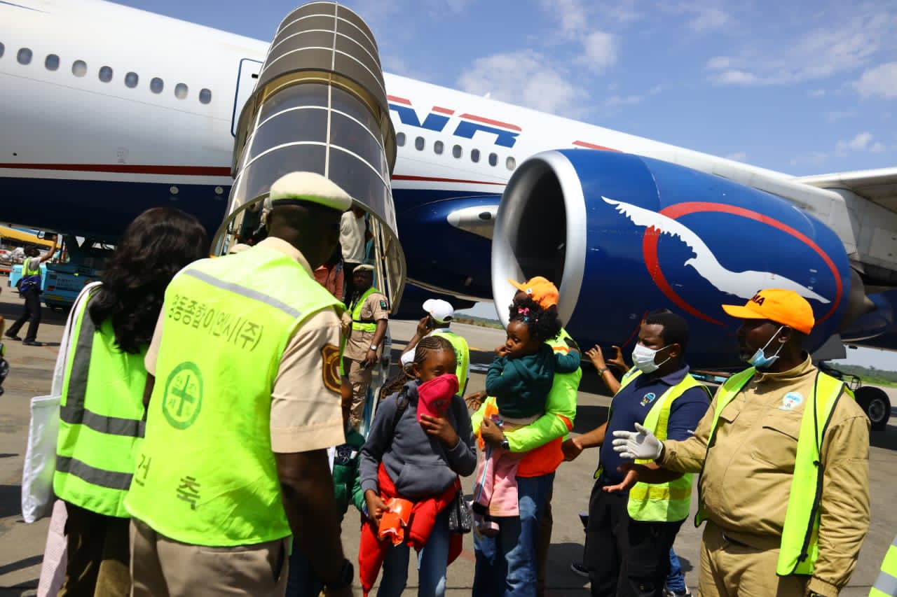 Ground crew in safety vests assist children at a jet bridge beside a large white and blue airplane with a prominent engine.