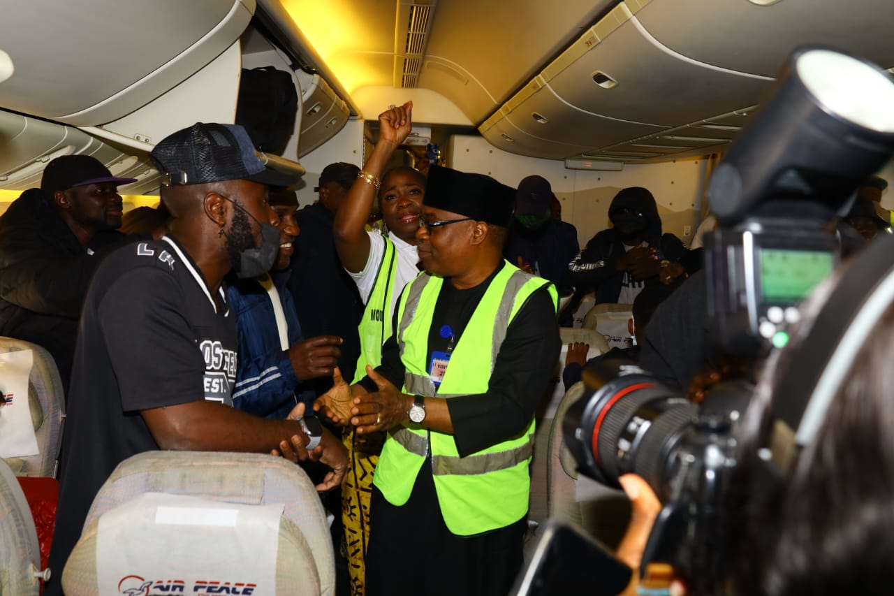 Crowded airplane cabin: a crew member in a neon safety vest speaks to standing passengers as a photographer films.