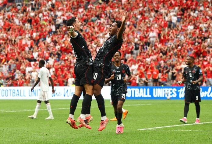 Canada Soccer players in black uniforms leap in celebration on the field as a packed crowd in red cheers behind them.