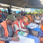THEWILL EDITORIAL: Malpractices Mar Ekiti Gov Poll Group of people in orange vests seated at blue tables under a tent, filling out forms for an election event.
