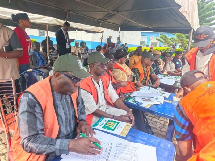 Group of people in orange vests seated at blue tables under a tent, filling out forms for an election event.