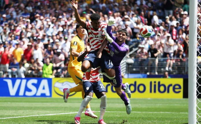 Soccer players contest the ball near the goal as a purple-clad goalkeeper dives to make a save, with a packed stadium behind.
