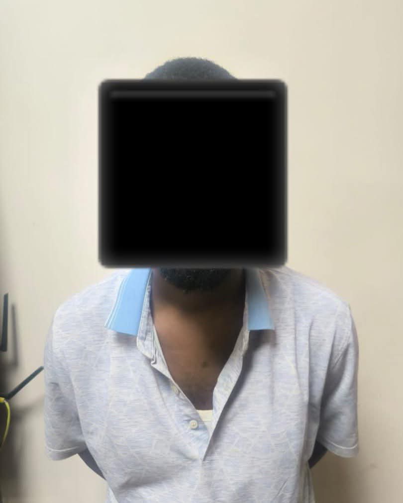 Person with face covered by a black square, wearing a light blue polo shirt, standing against a beige wall.
