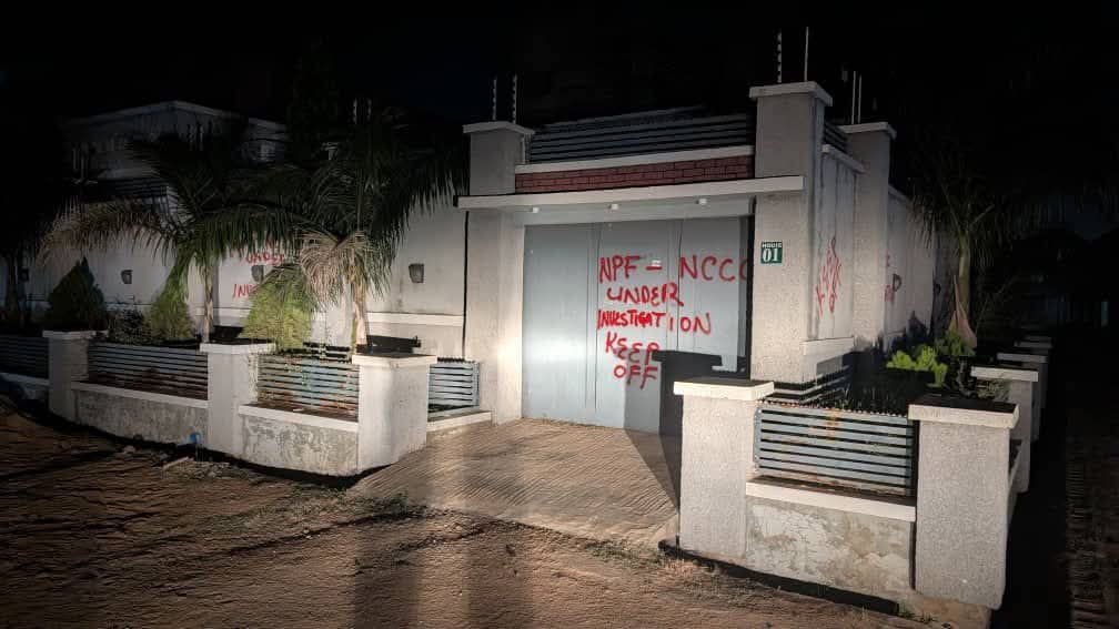 Nighttime view of a gated house with graffiti on the central metal door and palm trees in the yard.