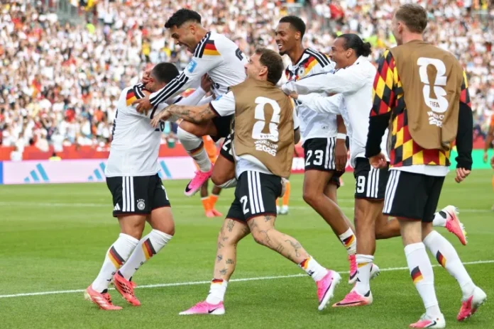 German national team players celebrate a goal on the soccer pitch, hugging and lifting a teammate as fans cheer in the background.