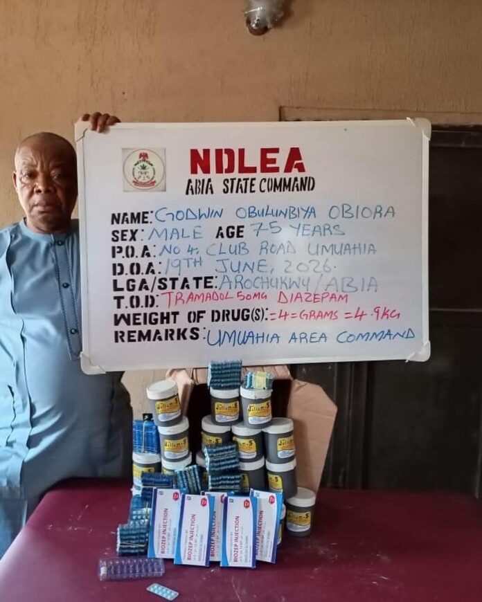 Godfrey Orji, 84, and Godwin Obulunbiya Obiora, 75, Man standing beside a board reading NDLEA Abia State Command, with a drug haul and packets arranged on a table in front of him.