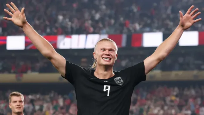 Haaland Soccer player with blond hair and a black jersey with number 9 raises arms in celebration on the field, crowd cheering in the stands.
