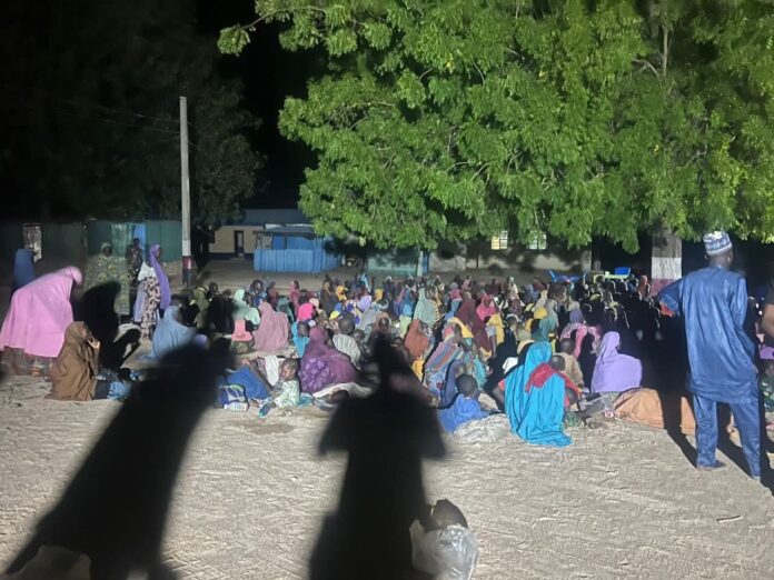IMG_6581 Large crowd seated on the dusty ground at night under a big tree, colorful clothing, likely a community gathering or meeting.