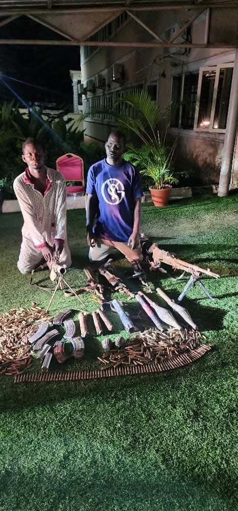 IMG_6586 Two men kneel beside a display of rifles, magazines, and ammunition arranged on the grass at night.