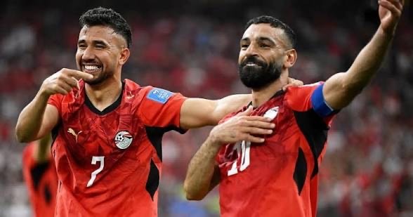Two football players in red jerseys celebrate on the field, arms around each other after a goal on the pitch (numbers 7 and 20 visible).