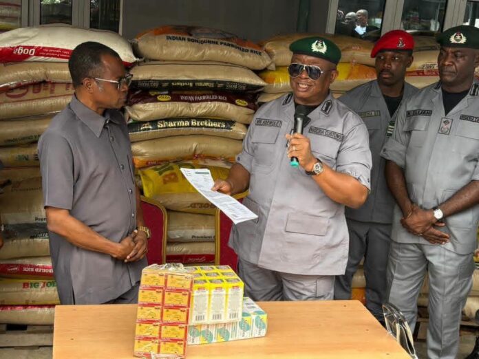 Uniformed officer speaks into a microphone beside stacked food boxes and rice sacks at a distribution event.