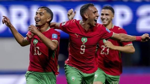 Three Morocco football players celebrate a goal, wearing red jerseys and green shorts, arms raised in jubilation on the field with numbers 11, 9, and 23 visible.