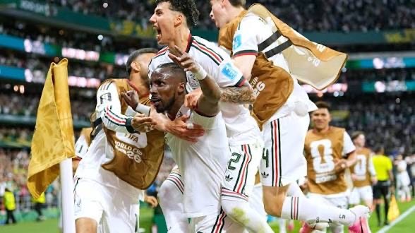 Group of soccer players in white uniforms celebrate a goal on the field, arms raised and shouting.