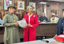 INEC Presents Certificates Of Return To Winners Of June 20 Bye-Elections A formal award ceremony in a council chamber: a man in olive-green traditional attire receives a certificate from a woman in a red suit; several officials watch from behind a long table.