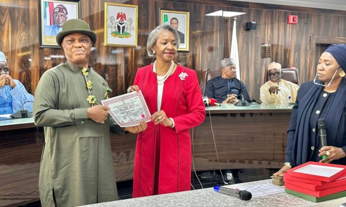 INEC Presents Certificates Of Return To Winners Of June 20 Bye-Elections A formal award ceremony in a council chamber: a man in olive-green traditional attire receives a certificate from a woman in a red suit; several officials watch from behind a long table.