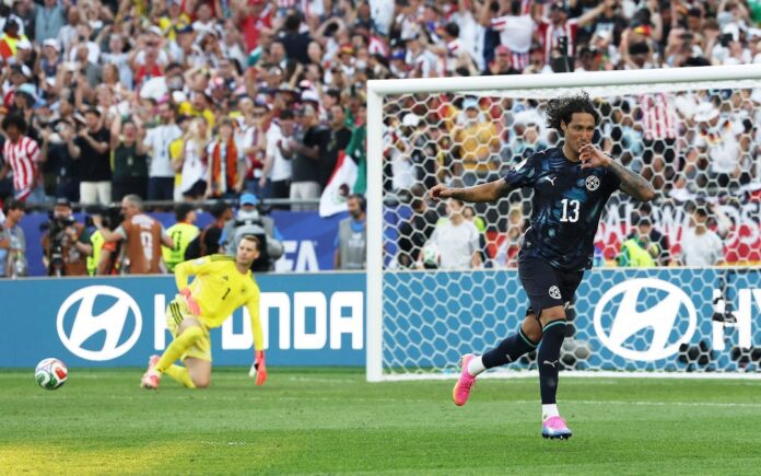Soccer scene: a player in a dark blue jersey #13 celebrates near the goal as the yellow-clad goalkeeper kneels on the pitch; a full stands crowd cheers behind them.