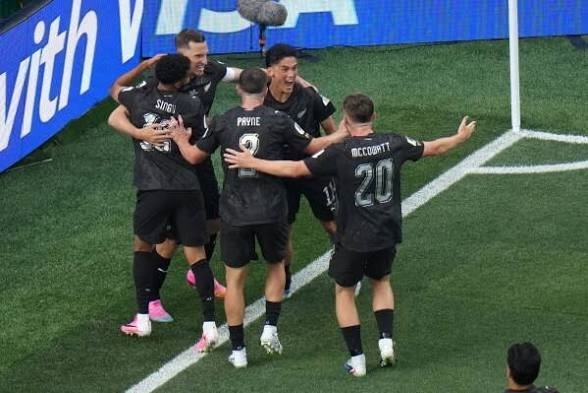 Group of five soccer players in black jerseys celebrate with arms around each other near the corner of the field after a goal.