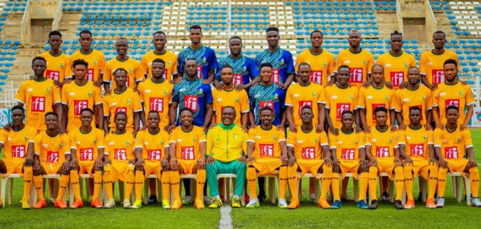 Group photo of a soccer team in yellow kits with red sponsor logos, posed on the field with blue and white stadium seats in the background. A coach sits center front in green and yellow.