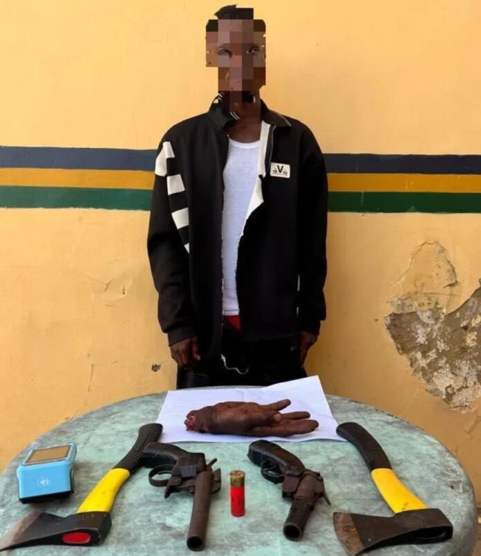 Lagos Police Nab 21-Year-Old With Human Body Part, Firearms, POS Machine, ATM Cards Man with a pixelated face stands behind a table displaying a severed hand placed on paper, with axes and other tools laid out.