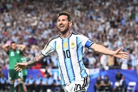 Soccer player in Argentina kit with number 10 celebrates a goal on the field, arms outstretched in a crowded stadium.