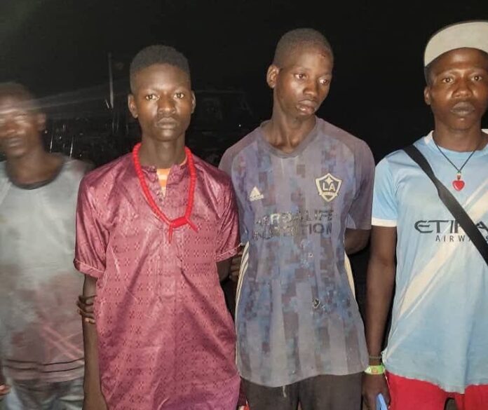 Four young men posing together at night, wearing casual outfits; one in a maroon tunic with a red bead necklace, others in a blue camo shirt and a light blue jersey with a cap.