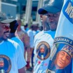 MUSAWAH: Musa Support Group Coordinator Hails Minister’s Confidence In Youths Two men in white shirts with circular logos chat at an outdoor gathering; one holds a flag showing a portrait.