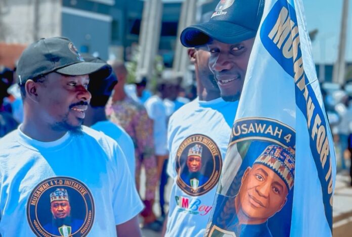Musa Support Group Two men in white shirts with circular logos chat at an outdoor gathering; one holds a flag showing a portrait.