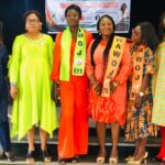 NAWOJ Recommits To More Advocacy, Inaugurates Bayelsa Chapter Six women stand on a stage in bright outfits, wearing sashes that read NAWOJ BYS, posing for a photo at an inauguration event with a banner in the background.