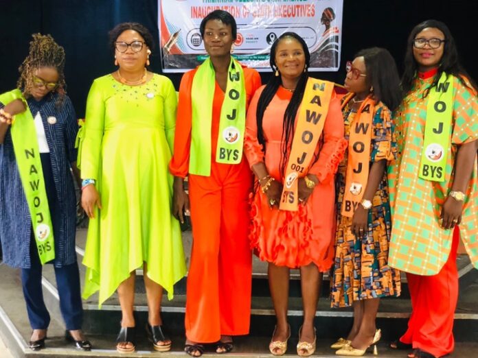 NAWOJ Six women stand on a stage in bright outfits, wearing sashes that read NAWOJ BYS, posing for a photo at an inauguration event with a banner in the background.