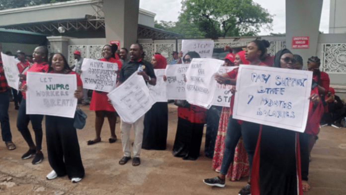 NUPRC-workers-e1690968913983 Group of protesters, mostly women in red and black clothing, standing outside a building and holding signs calling for higher pay and better conditions for staff.