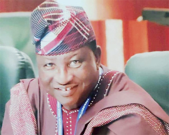 Otunba-Oladiti Smiling man in traditional Nigerian attire with a patterned cap and maroon outfit, seated indoors with a blue lanyard visible.