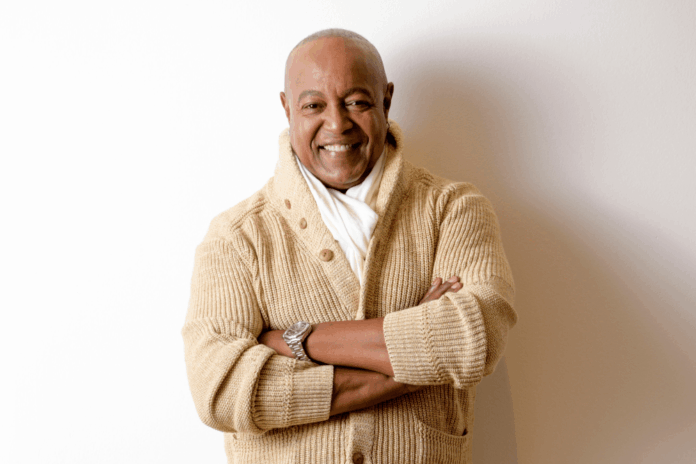 Smiling man in a beige cardigan with arms crossed, standing against a white wall.