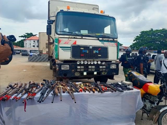 Police Intercept Military Uniforms, Narcotics In Lagos, Table displays seized weapons—rifles, pistols, and knives—laid out in front of a large truck, with police officers and bystanders nearby.