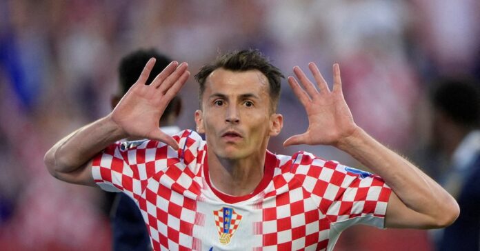 RS2GUKMJCFNJNKN7CY2HPTVJ3I Croatian soccer player in a red-and-white checker kit celebrates with hands near his ears on the field.