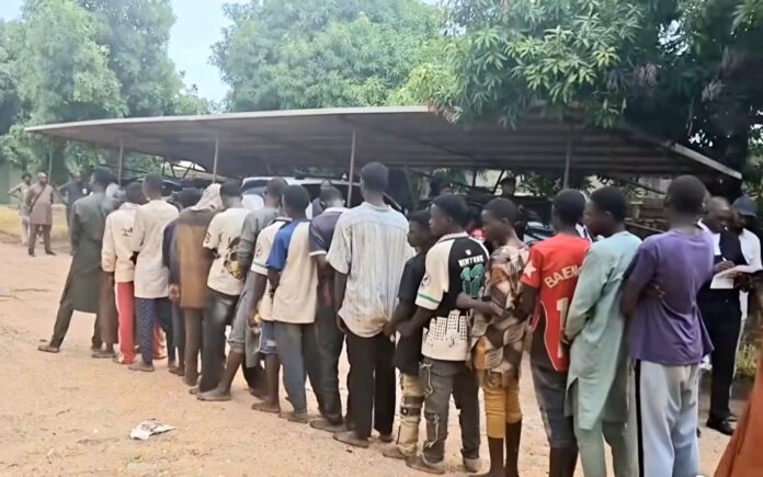A long line of people standing in a queue under a metal carport-style shelter in an outdoor, dusty area with trees in the background.
