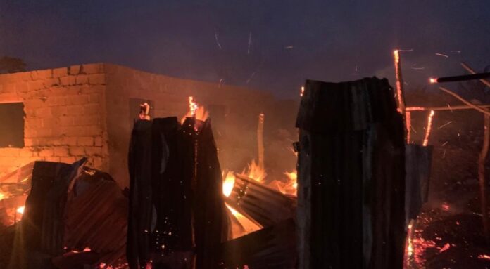 Nighttime scene of burning wooden debris and a brick wall, with large flames and glowing embers.