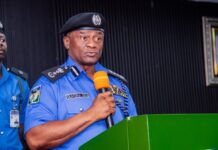 Your Jobs Are Safe, State Police Won’t Scrap Federal Force – IGP Disu Assures Personnel Police officer in blue uniform speaking into an orange microphone at a green podium, delivering a formal address.