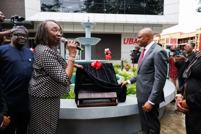 UBA-unveils-in-UNILAG A woman in a black-and-white polka-dot outfit speaks into a microphone beside a covered plaque at a formal outdoor ceremony, while a suited man watches nearby.