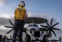 US Hits Iran In Fresh Strikes, Targets Missiles, Drone Storage Ground crew member in a yellow safety vest walks toward a twin-propeller cargo plane on a runway.