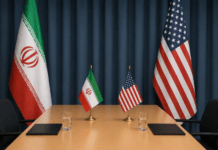 Explosions Shake Southern Iran as U.S. Escalates Military Campaign Diplomatic conference table with Iranian and American flags, two small flags, notebooks, and water glasses against a dark curtain backdrop.