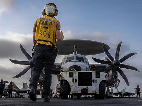 US Ground crew member in a yellow safety vest walks toward a twin-propeller cargo plane on a runway.