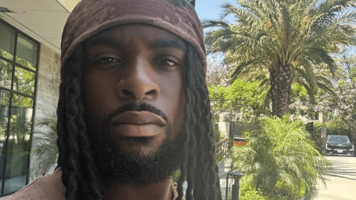 british-nigerian-singer-songwriter-talay-riley Close-up portrait of a man with long dreadlocks and a brown head wrap, outdoors with palm trees and a parked car in the background.