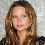 Lilo & Stitch Star Daveigh Chase Dies At 35 Portrait of a young woman with long wavy light brown hair and blue eyes, wearing a black lace top against a gray textured background, looking to the side.