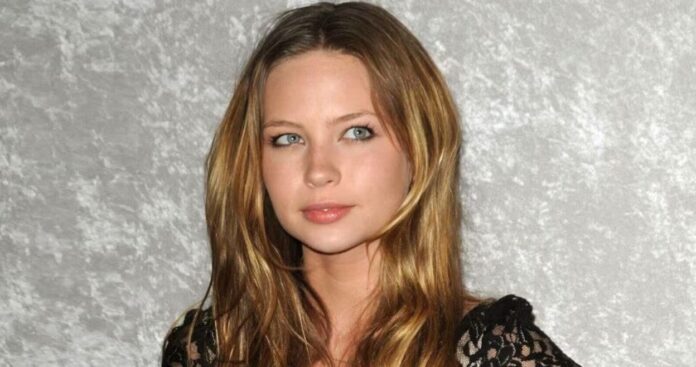 Portrait of a young woman with long wavy light brown hair and blue eyes, wearing a black lace top against a gray textured background, looking to the side.