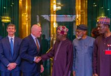 Tinubu Courts Mastercard Investment, Says Nigeria’s Youths Are Nation’s Greatest Asset Two men in business suits shake hands while a group of men in traditional attire look on in a gold-and-glass lobby setting; formal greeting scene.