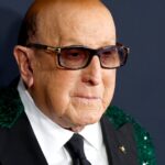 Legendary Music Executive Clive Davis Laid To Rest As Music Stars Celebrate Legacy Older man wearing dark sunglasses and a green sequined blazer at a formal event, neutral backdrop with logos behind him