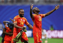 Historic DR Congo Roar Into Knockouts After Stunning Comeback Against Uzbekistan Two male soccer players in red uniforms celebrate on the field, with arms raised.