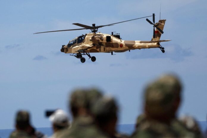 None Desert camouflaged military helicopter in flight above a group of blurred soldiers on the ground against a blue sky.
