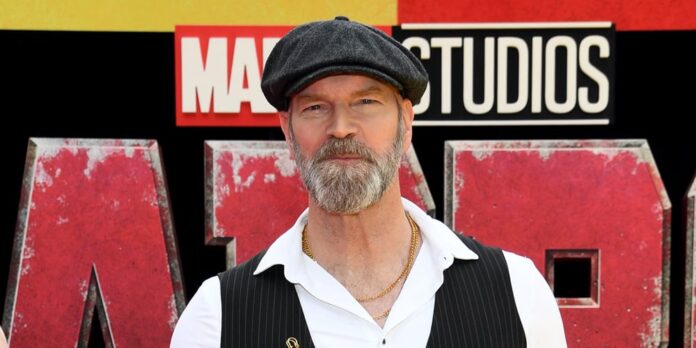 Man with a gray beard wearing a black beret and vest poses in front of a Marvel Studios backdrop at a red-carpet event.